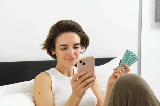 Portrait of smiling young woman reading on mobile phone, searching medication dosage instruction, her prescription on medical app, holding pills, sitting on bed.