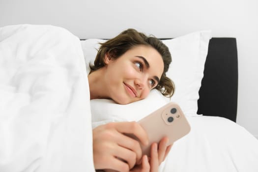 Portrait of woman lying in her bed, checking phone, sets up alarm clock to wake up in the morning.