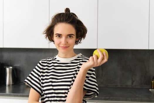 Portrait of young female model, posing in the kitchen, holding an apple, smiling and looking at camera.