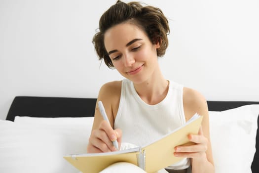 Portrait of woman sitting in bed with journal, writing in her diary, thoughts or feelings in the morning, holding notebook and smiling.