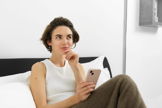 Thinking woman in bedroom, holding smartphone, making decision, placing an order, online shopping, spending time and relaxing in bed.