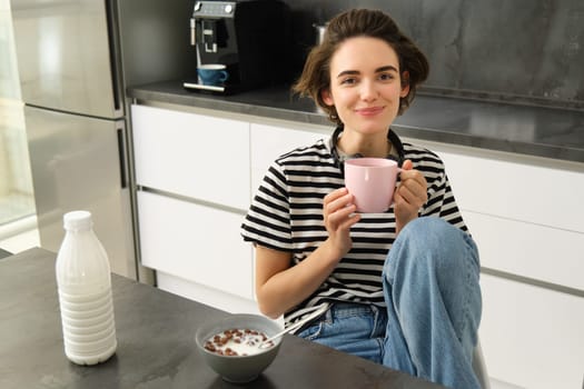 Cute brunette woman, student eating breakfast in morning, drinking hot tea, smiling and looking pleased.