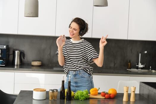 Portrait of happy girl cooking salad and listening to music in headphones, dancing while making a meal in the kitchen.