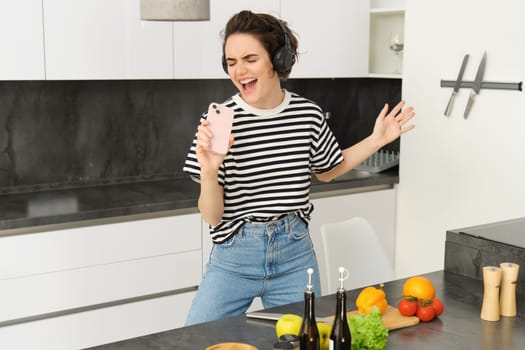 Young woman cooking meal in kitchen, listening to music in wireless headphones, singing into smartphone microphone, making salad, chopping vegetables.