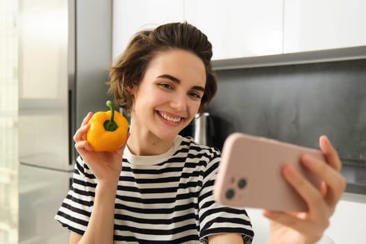 Smiling, beautiful young woman taking selfie with fresh yellow pepper, food blogger taking selfie with vegetables in the kitchen, vegan making a meal and recording video for social media.
