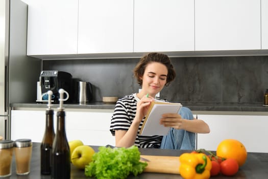 Portrait of young smiling woman in kitchen, holding notebook, making notes for recipe, writing grocery list, cooking salad, sitting near vegetables and chopping board.