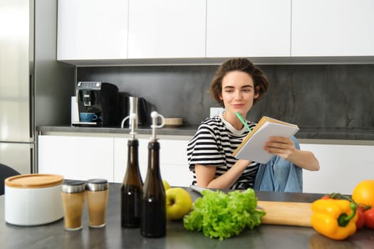 Portrait of young smiling woman in kitchen, holding notebook, making notes for recipe, writing grocery list, cooking salad, sitting near vegetables and chopping board.
