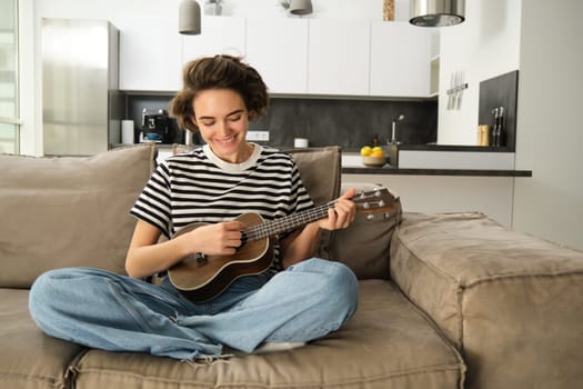 Portrait of cheerful young woman playing her ukulele, singing and laughing, sitting in living room at home. Lifestyle and music concept