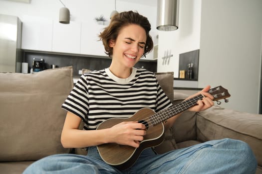 Portrait of young modern woman, student playing ukulele at home, sitting with small guitar, singing and feeling happy, sitting on sofa. Lifestyle and music concept