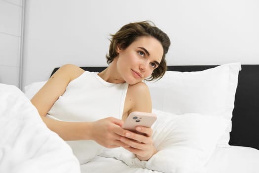 Portrait of woman in bed, lying on white sheets, using smartphone, holding mobile phone. Lifestyle and technology concept