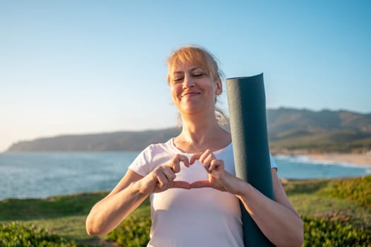 Mature woman with yoga mat on coast. Adult woman on nature smiling on green seashore standing with yoga mat and shows heart gesture