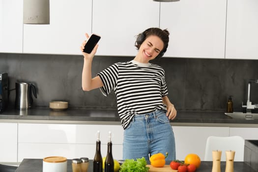 Portrait of happy dancing woman, cooking in the kitchen and listening music in headphones, chopping vegetables for salad and singing with carefree face.