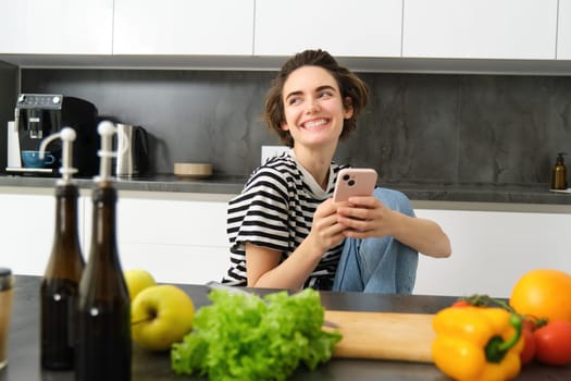 Portrait of young woman smiling, laughing and using smartphone while cooking, searching recipes on smartphone app, preparing meal from vegetables in her kitchen at home.