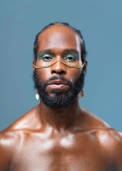 Black gay stands isolated on blue background and stares into camera with serious expression on face. Close-up portrait of handsome adult transgender with blue eyeshadows and golden accessory on face