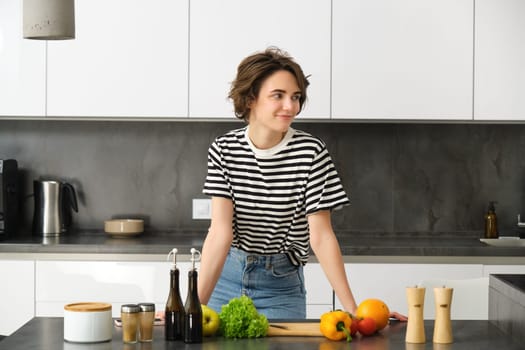 Portrait of smiling cute young woman making breakfast, chopping vegetables in the kitchen, preparing vegan meal.