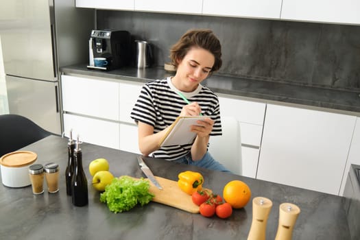 Portrait of woman writing down her recipe, making notes in notebook during cooking, sitting in the kitchen with chopping board and vegetables, preparing a meal.