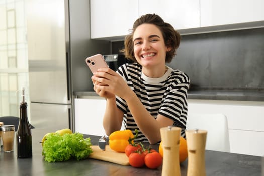 Portrait of brunette girl cooking food in the kitchen, searching recipes on social media app, holding mobile phone, standing near chopping board and vegetables, preparing healthy vegetarian meal.