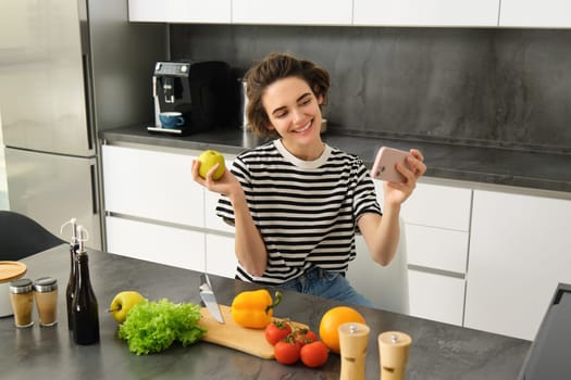 Smiling young woman, food blogger recording a video on smartphone with food, cooking and making selfie, taking pictures during meal preparation, sitting near vegetables and holding an apple.