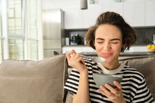 Portrait of young female model, student eating quick breakfast, holding bowl of granola or cereals with milk, sitting on sofa in living room.