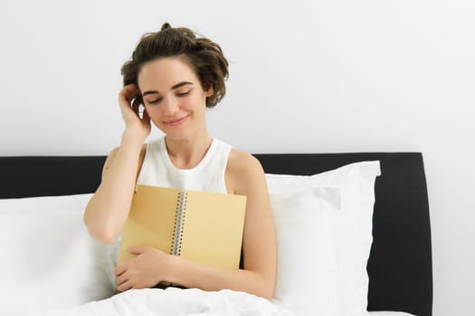 Portrait of beautiful woman in bed, sitting with her journal, writing in diary and smiling, tucking hair behind ear, reading notebook. Lifestyle and people concept