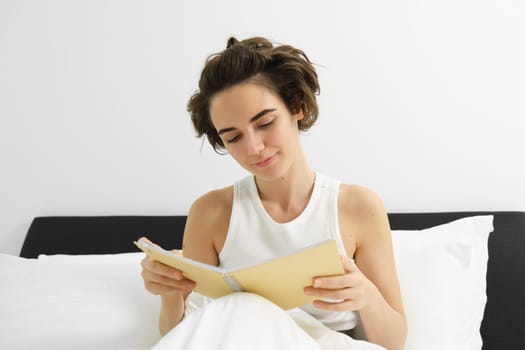 Close up portrait of young woman, reading journal, sitting in bed and studying, revising for exam before going to sleep.