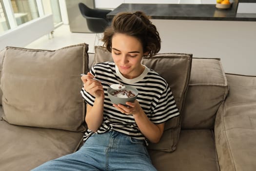 Portrait of young female model, student eating quick breakfast, holding bowl of granola or cereals with milk, sitting on sofa in living room.