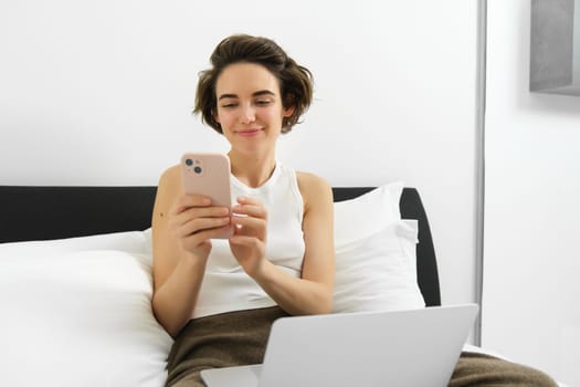 Work from home concept. Young woman sitting on bed, freelancing, working on laptop, using smartphone, looking at mobile phone with pleased smiling face.