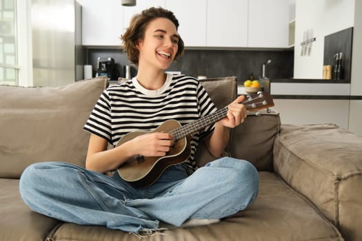 Cheerful young woman with ukulele, playing musical instrument, holding small guitar and singing, sitting on sofa on crossed legs, resting in living room.