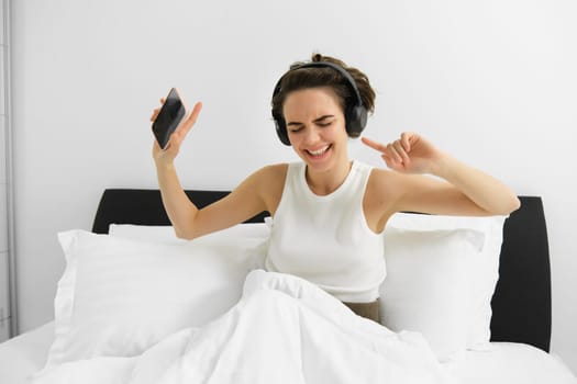 Portrait of happy young woman in bed, listening to music in wireless headphones, dancing in her bedroom, holding mobile phone in hand. Copy space