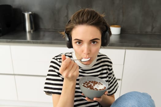 Beautiful woman, stylish girl eating cereals with milk, holding spoon near mouth and bowl in hands, having her breakfast in the morning, sitting in kitchen, listening music in headphones.