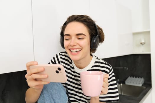 Lifestyle concept. Young modern woman watches movie on smartphone, wearing headphones, drinks coffee or tea in the kitchen.