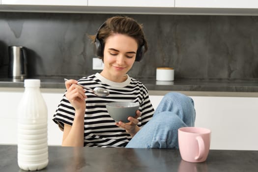 Portrait of smiling young candid woman, student eating morning cereals, having her breakfast, listening music in wireless headphones, sitting in the kitchen.