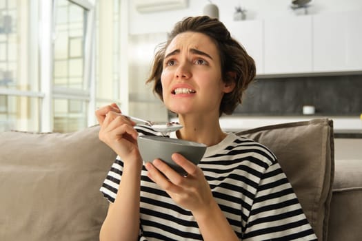 Image of woman eating breakfast in front of tv, looking at screen with worried face, holding bowl of cereals with milk and a spoon, spending time in living room.