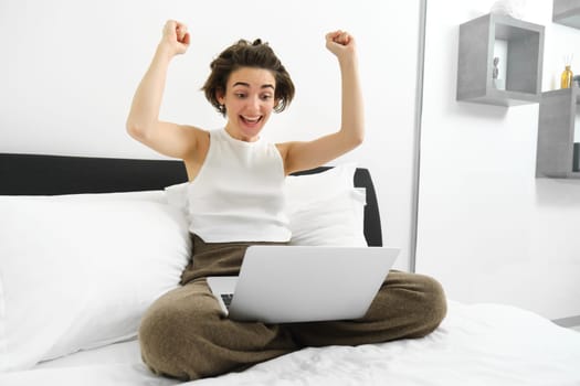 Winning girl, looking at laptop with hands raised up, sitting on bed, watching something in laptop and triumphing, celebrating victory.