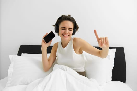 Image of beautiful smiling woman, dancing on bed, waking up and listening to music in wireless headphones, energizing, happy morning. Lifestyle concept