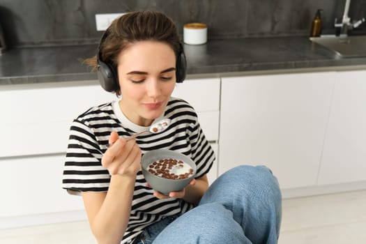 Happy brunette woman in the kitchen, eating cereals with milk for breakfast, listening music in headphones, having her morning meal.
