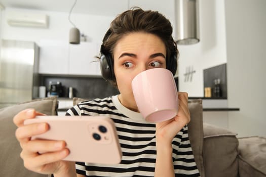 Portrait of girl sipping tea and watching tv show on smartphone with interest, stating at mobile phone amazed, sitting on sofa in living room.