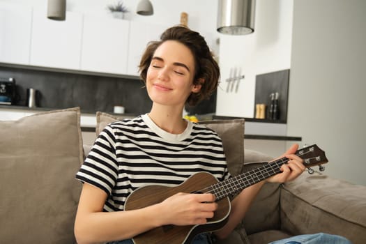 Close up portrait of beautiful smiling woman, playing ukulele, strumming strings with closed eyes and pleased face, sitting in living room.