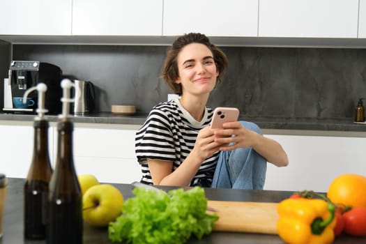 Portrait of brunette young woman with smartphone, sitting in the kitchen, watching cooking recipe videos, has chopping board and vegetables.