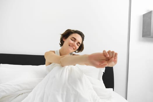 Portrait of smiling young woman wakes up in her bed, sitting under white linen sheets with closed eyes and stretching her hands with satisfaction.