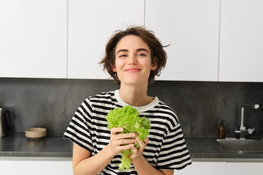 Portrait of happy and healthy young woman, following her diet, posing with lettuce leaf and smiling, cooking in the kitchen, vegetarian loves her vegetables.