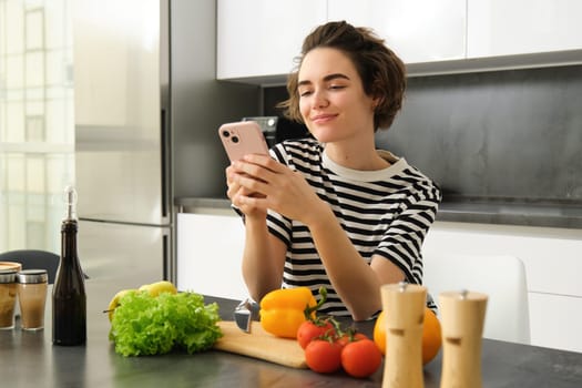 Portrait of young woman food blogger, posting on social media recipe, vegan salad, standing in the kitchen with smartphone, chopping vegetables.