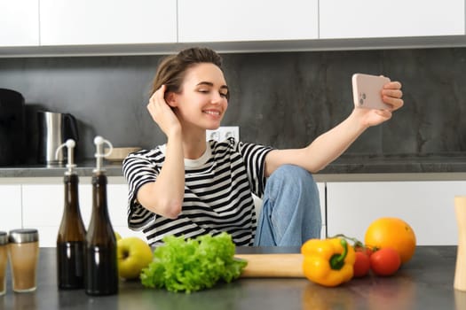 Cute young woman, modern lifestyle blogger, taking selfie while cooking salad in the kitchen, using smartphone to make photos for social media app.