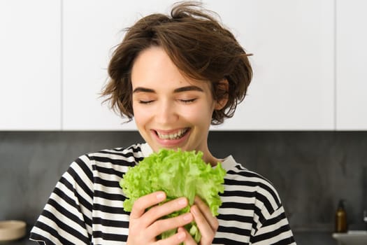 Portrait of happy and healthy young woman, following her diet, posing with lettuce leaf and smiling, cooking in the kitchen, vegetarian loves her vegetables.