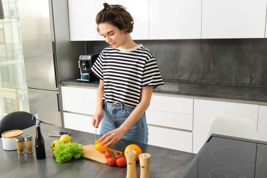 Portrait of modern young woman standing in the kitchen with vegetables, chopping them on counter, holding knife, cooking meal, vegetarian food, concept of healthy lifestyle an diet.