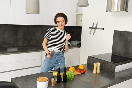 Modern happy woman dancing and cooking, listening music in headphones, making a vegan healthy meal, vegetarian food.