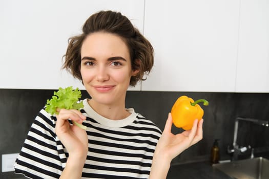 Close up of cute smiling woman, showing lettuce leaf and yellow pepper, cooking salad in the kitchen, likes eating vegetables as healthy snack.