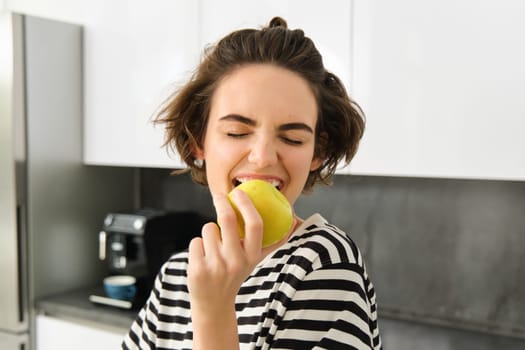 Close up portrait of young brunette woman biting an apple with pleasure, has pleased smile on her face, standing in the kitchen, having healthy snack for lunch, eating fruits.