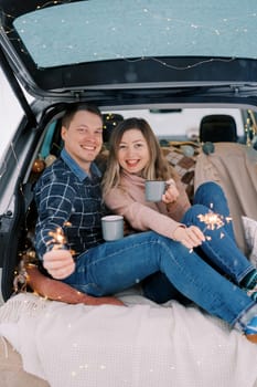 Smiling couple with mugs of coffee and sparklers in their hands are sitting on bedspreads in the trunk of a car. High quality photo