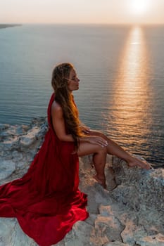 Woman sunset sea red dress, side view a happy beautiful sensual woman in a red long dress posing on a rock high above the sea on sunset